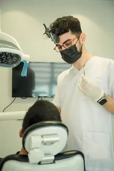 Médico em pé de óculos de grau, máscara, luva e pacoente sentado em cadeira com tv ao funso