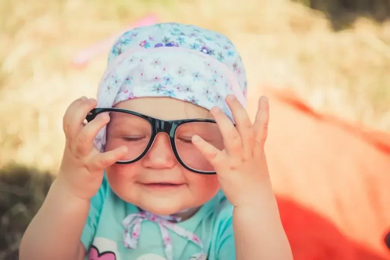 Bebe de toca e óculos de grau, com olhos fechados e mãos na frente do óculos